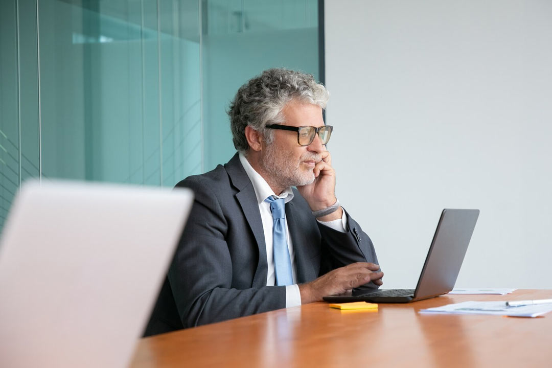 Contador experiente, com os cabelos brancos, óculos de grau, terno e gravata, sentado em mesa de escritório com cara de preocupação analisando dados em um notebook
