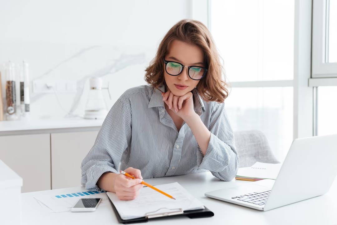 Jovem empresária usando óculos com os cabelos castanhos claros soltos, lisos, na altura do ombro, analisa informações em uma prancheta, sentada em uma mesa de escritório, com o notebook aberto