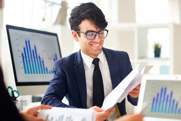 Jovem profissional de contabilidade sorrindo e vestindo roupa social, analisa informações, sobre a empresa, entre as telas de dois computadores com a imagem de gráficos