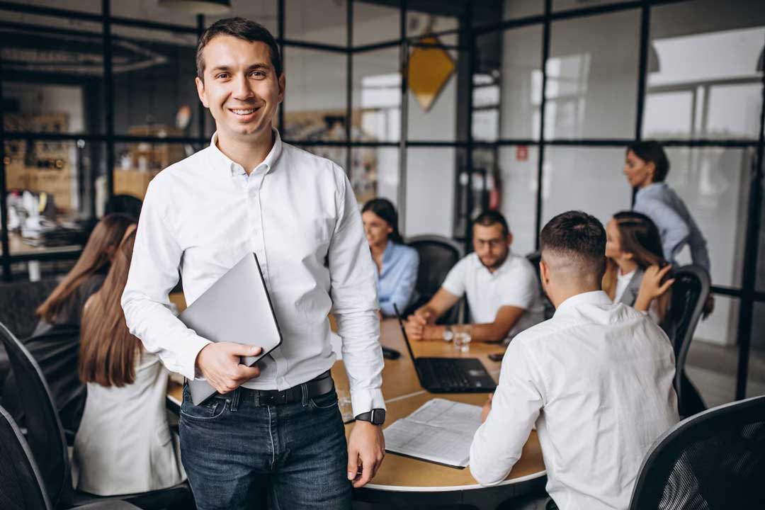 Jovem empresário sorrindo, segurando um notebook a frente de uma mesa com um grupo de empresários sentados com caderno de anotação e notebook, em reunião