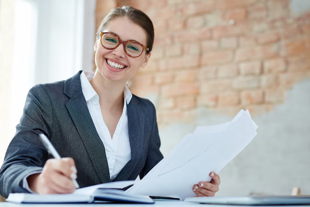 Jovem mulher profissional de contabilidade, loira de cabelos presos, sorrindo, segurando folhas com informações da empresa e anotando dados