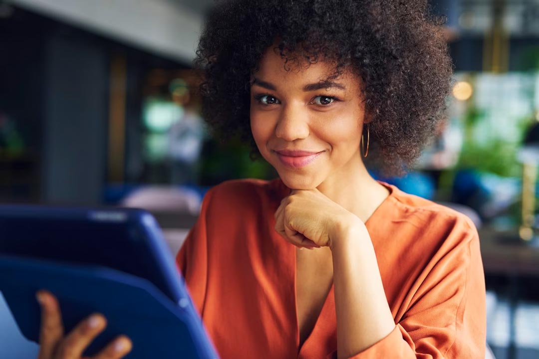 Jovem mulher negra, com blusa cor de laranja, segurando um tablet e sorrindo com a mão no queixo dentro do escritório