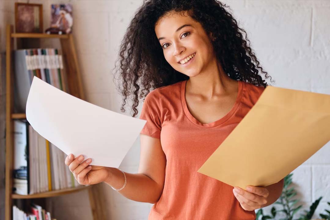 Jovem mulher negra, sorrindo, segura em uma das mãos um envelope laranja e na outra mão um documento impresso em folha branca