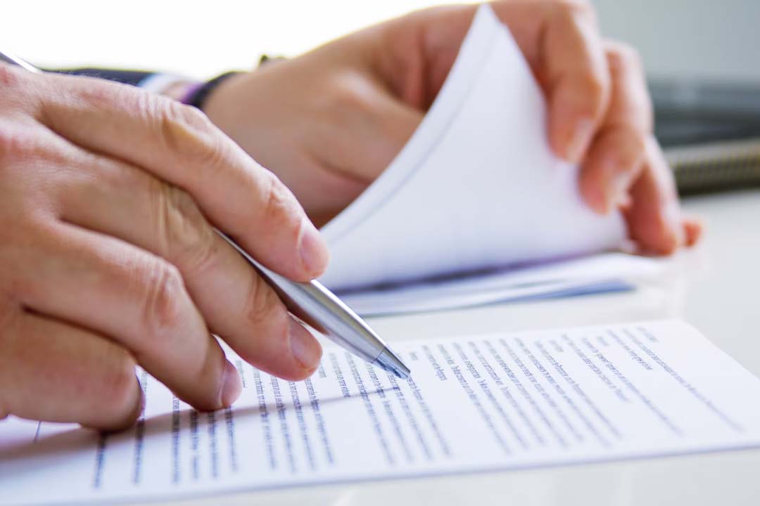 Mãos de um executivo analisando e fazendo pontuações sobre um documento que esta em cima de uma mesa branca