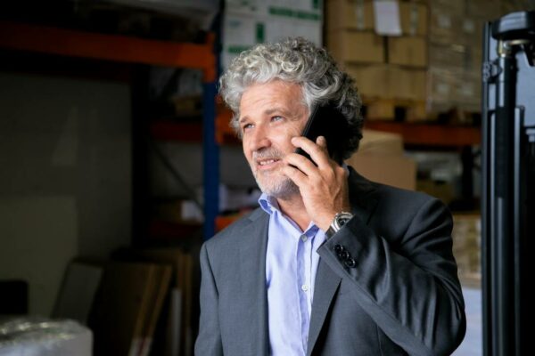 empreendedor-maduro-positivo-pensativo-em-pe-no-armazem-e-falando-no-celular-prateleiras